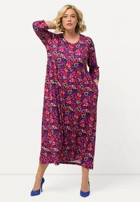 Langes lila Kleid mit lebhaften geometrischen Mustern in Pink, Rot und Creme. Verfügt über einen runden Ausschnitt und Taschen. Stilisiert mit blauen Absätzen.