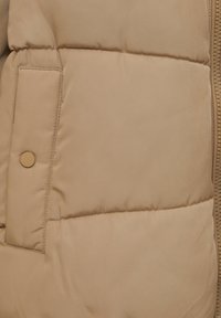 Chaqueta acolchada beige con textura acolchada, bolsillo con botones y cremallera visible. La tela parece suave y ligera.