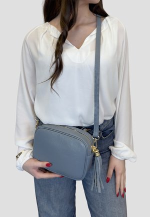 Kvinde iført hvid bluse og blå jeans, der holder en lille grå lædertaske med tværrem, pyntet med kvast og guldsmedede detaljer.