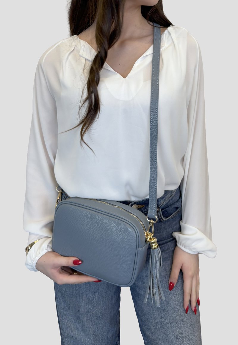 Kobieta ubrana w białą bluzkę i niebieskie jeansy, trzymająca małą szarą skórzaną torebkę na ramię z frędzlami i złotymi ozdobami.