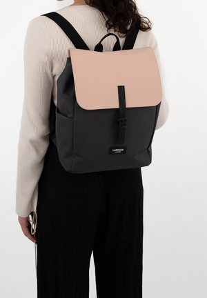 Černý a růžový batoh s hladkým, strukturovaným designem; má nastavitelné popruhy a přední zapínání na přezku. Viditelná značka značky.