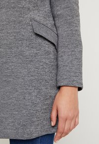 Harmaa, teksturoitu takki, jossa on käännetty hihansuu ja sivutasku. Kangas näyttää pehmeältä ja hieman ribbineulotulta, ja takissa on rento muotoilu.