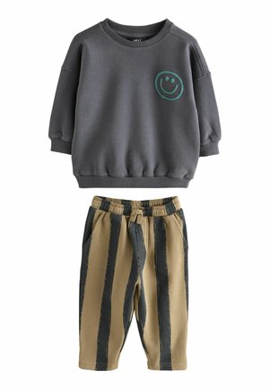 Sweatshirt gris charbon à manches longues avec un visage souriant vert et pantalon beige avec de larges rayures verticales noires et taille élastique.