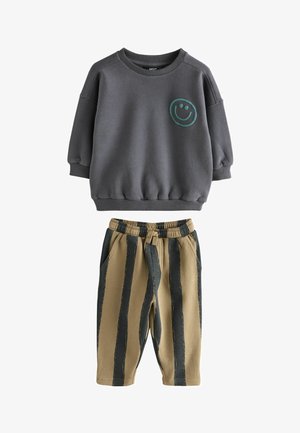 Koolgrijze sweatshirt met lange mouwen en een groene glimlachende emoji, gecombineerd met beige broek met brede verticale zwarte strepen en een elastieken tailleband.