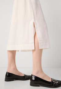 Gambe inferiori che indossano mocassini neri in pelle lucida con dettagli in metallo, abbinati a pantaloni bianchi corti a gamba larga con fessure laterali.