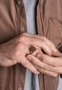 Gold metal ring with a hexagonal brown wooden inlay, featuring a cutout design on one side, worn on a hand against a textured brown shirt.