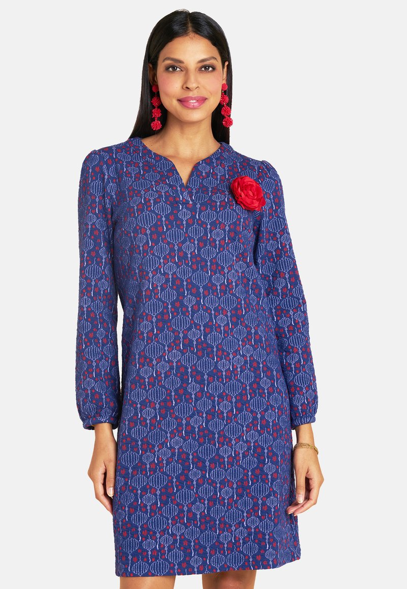 Blau gemustertes Kleid mit langen Ärmeln, das ein florales Design und eine rote Blütenakzent auf der Brust aufweist. Aus leichtem Stoff gefertigt.