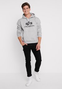 Siva dukserica s kapuljačom s crnim logom na kojem piše "Alpha Industries." Ima kapuljaču s vezicama, rebraste manžete i prednji džep. Uparena s crnim hlačama.