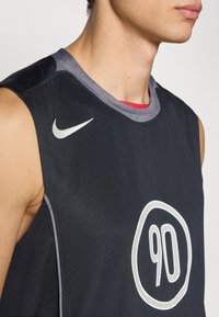 Mustapohjainen urheilupaita ilman hihoja, jossa on harmaat yksityiskohdat. Paidassa on suuri valkoinen pyöreä graafinen kuva, jossa on numero '90' ja hopeanvärinen Nike-logo.