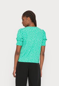 Top verde com bolinhas, mangas curtas e volumosas, tecido texturizado e ajuste descontraído. Usado com parte de baixo preta, mostrando uma vista traseira suave.