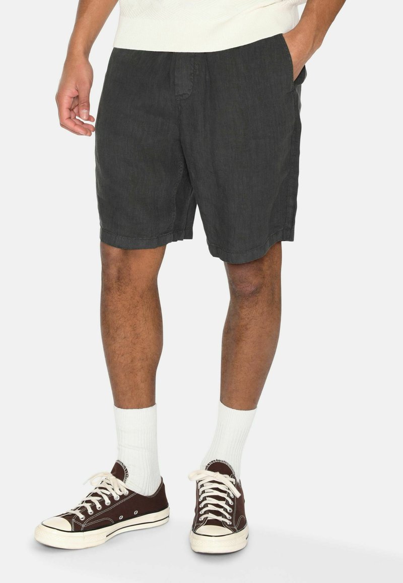 Dunkelgraue Leinen-Shorts mit lockerer Passform, seitlichen Taschen und flacher Vorderseite, kombiniert mit weißen Socken und braunen Leinenschuhen.