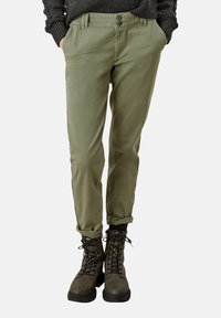Olivově zelené chinos kalhoty s hladkou texturou, slim fit a ohrnutými nohavicemi, spárované s tmavě hnědými šněrovacími botami s robustními podešvemi.