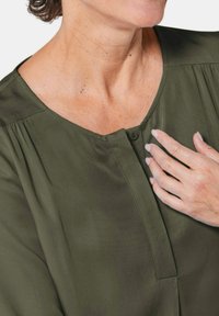 Olivengrüne Bluse mit rundem Ausschnitt, Knopfverschluss und gefalteten Details an den Schultern. Weicher Stoff und lockere Passform.
