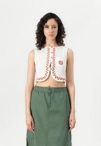 Korte witte top met oranje geometrische borduursels, decoratieve knopen en een sleutelgaatje, gecombineerd met een groene rok met elastische taille.