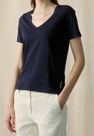 Frau trägt ein marineblaues kurzärmeliges V-Ausschnitt-Shirt und cremefarbene Hose, eine Hand in der Tasche vor einem einfarbigen Hintergrund.