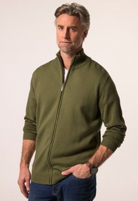 Muž se šedými vlasy a vousy, oblečený v olivově zeleném svetru na zip, modrých džínách a černých náramkových hodinkách, stojí s jednou rukou v kapse.