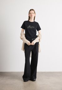 Sort t-shirt med et struktureret sølvdesign, parret med løse sorte bukser og et lys beige yderlag draperet over armene.