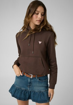 Donna che indossa una felpa marrone con coulisse e un piccolo logo, abbinata a una minigonna in denim con volant e una cintura marrone.