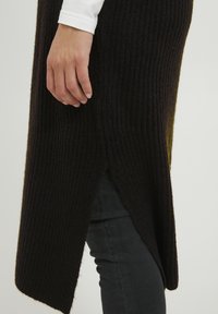 Pull en tricot noir avec texture côtelée, présentant une fente sur le côté pour plus de liberté de mouvement, porté sur un jean foncé et un haut à manches longues blanc.