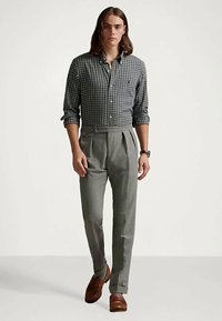 Chemise boutonnée à carreaux gris, pantalons plissés gris, chaussures en cuir marron. Coupe ajustée avec les manches retroussées et un léger détail de montre au poignet.