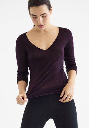 Femme portant un haut à manches longues violet foncé avec un col en V et un pantalon noir, tenant l'ourlet de son haut tout en regardant vers le bas.