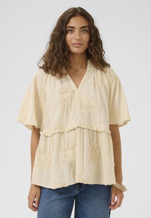 Mujer con cabello castaño ondulado, vestida con una blusa beige bordada con mangas acampanadas y jeans azules, sobre un fondo liso.