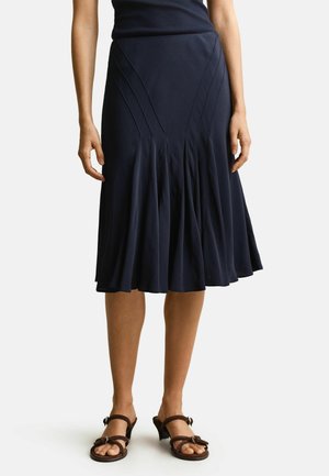 Frau trägt einen marineblauen, plissierten Midirock und braune Sandalen mit Absatz, steht vor einem schlichten hellen Hintergrund.