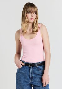 Giovane donna con capelli biondi che indossa un top rosa senza maniche, jeans blu e una cintura nera, in piedi su uno sfondo bianco semplice.