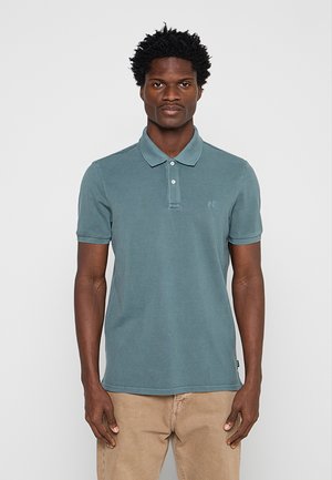 Uomo con capelli corti e ricci che indossa una polo turchese e pantaloni beige, in piedi davanti a uno sfondo grigio chiaro uniforme.