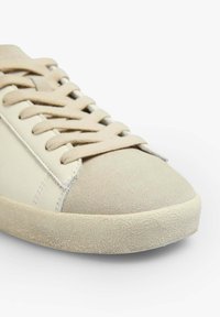 Basket beige avec un bout en daim texturé, des côtés en cuir lisse et des lacets plats. Elle présente une semelle en caoutchouc à motif avec un profil bas.