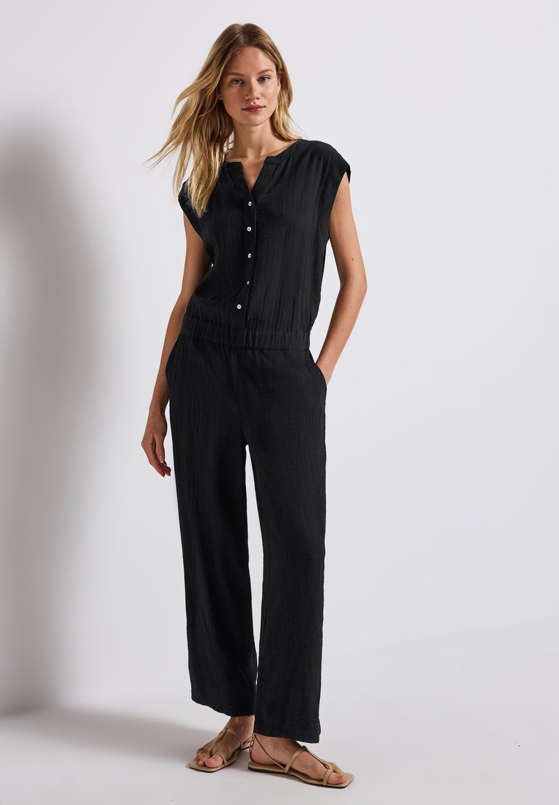 Zwarte jumpsuit met korte mouwen, knopen aan de voorkant, elastische taille en brede pijpen. Gemaakt van textuurstof, gecombineerd met platte sandalen.