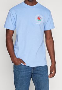 Světle modré bavlněné tričko s kulatým výstřihem, krátkými rukávy a potištěným designem růže na levé hrudi s textem: "Oddělení vizuálního designu."