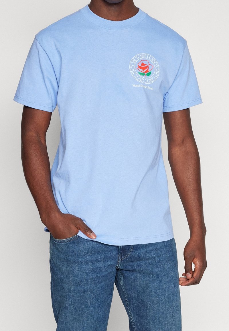 Světle modré bavlněné tričko s kulatým výstřihem, krátkými rukávy a potištěným designem růže na levé hrudi s textem: "Oddělení vizuálního designu."