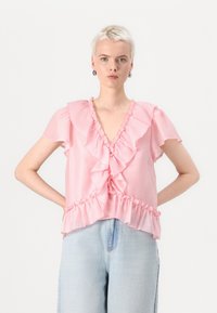 Blusa rosa con volant, maniche corte, scollatura a V profonda e design a strati. Tessuto leggero con una consistenza morbida, abbinata a denim chiaro.