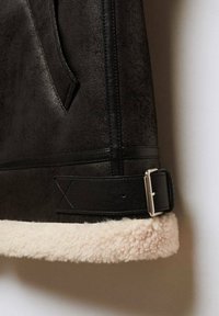Veste en daim marron avec accents en cuir noir, dotée d'une bordure en shearling beige douce et d'une boucle métallique sur la sangle réglable.