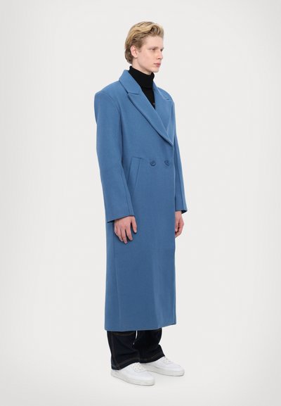 Modrá vlnená bunda s dvojradovým dizajnom, širokými klopami a bočnými vreckami, nosená cez čierny rolák a tmavé nohavice.