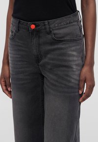 Person som har på seg falmede svarte jeans med fremtredende slitasje og en knallrød knapp, kombinert med en svart topp.