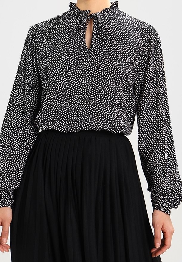 Blouse noire à manches longues avec pois blancs et col à volants, associée à une jupe plissée noire taille haute.