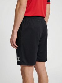 Bărbat purtând pantaloni scurți sport negri Hummel și un tricou roșu, stând lateral pe un fundal simplu și deschis.