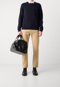 Marineblauer Pullover mit kleinem rotem Logo, beigefarbene Hose, schwarze Lederhandtasche in runder Form und schwarze Schnürschuhe, stehend vor weißem Hintergrund.