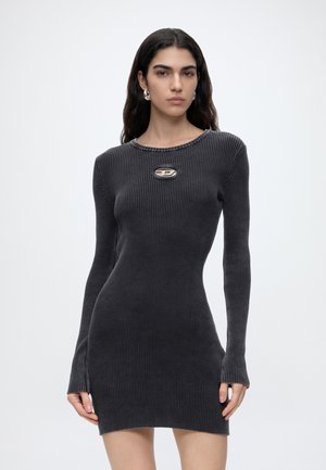 Femme aux longs cheveux foncés portant une robe côtelée gris foncé ajustée avec des manches longues et une petite découpe ovale près du col.