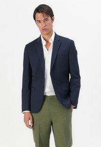 Marineblazer med to frontknapper, notched lapels og lommer; kombinert med en hvit skjorte og grønne bukser. Teksturert stoff.