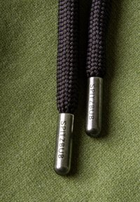 Black braided shoelaces with silver metal aglets engraved with "SPITZBUB," resting on a textured green fabric background.