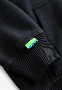 Felpa nera con orlo e polsini a coste. Presenta una piccola etichetta verde e blu con la scritta "by Superdry" sulla manica sinistra. Texture morbida.