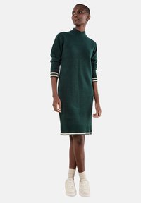 Abito verde lavorato a maglia con collo alto e polsini a coste. Accentuato da strisce bianche sui polsini e sull'orlo. Abbinato a sneakers bianche.