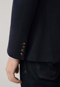 Marineblazer mit strukturiertem Finish, ausgestattet mit vier goldenen Knopfakzenten an den Manschetten, getragen über dunklen Jeans.