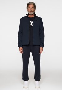 Merinovihreä kevyt takki korkealla kauluksella, mustalla vetoketjulla ja joustamalla hihnalla, yhdistettynä merinovihreään t-paitaan ja vastaaviin verkkareihin. Valkoiset lenkkarit.