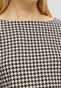 Schwarzes und weißes Hahnentritt-Muster auf einem glatten Stoff. Quadrat-Ausschnitt, weit geschnitten und minimalistische Nähdetails.