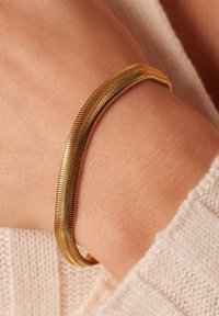 Gouden armband met een strakke, gladde vormgeving en geribbelde details, rustend op een pols tegen een lichtgekleurde gebreide trui.