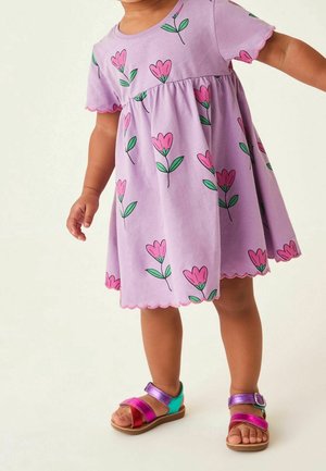 Robe lilas à manches courtes, motif floral rose et vert, ourlet festonné. Portée avec des sandales multicolores aux brides roses, violettes et vertes.